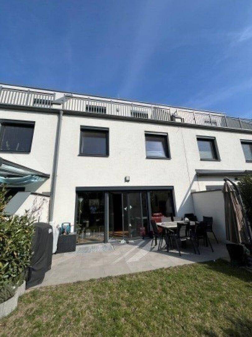 Moderne Hausfassade mit großer Glasfront, Terrasse und kleinem Gartenbereich unter strahlend blauem Himmel.