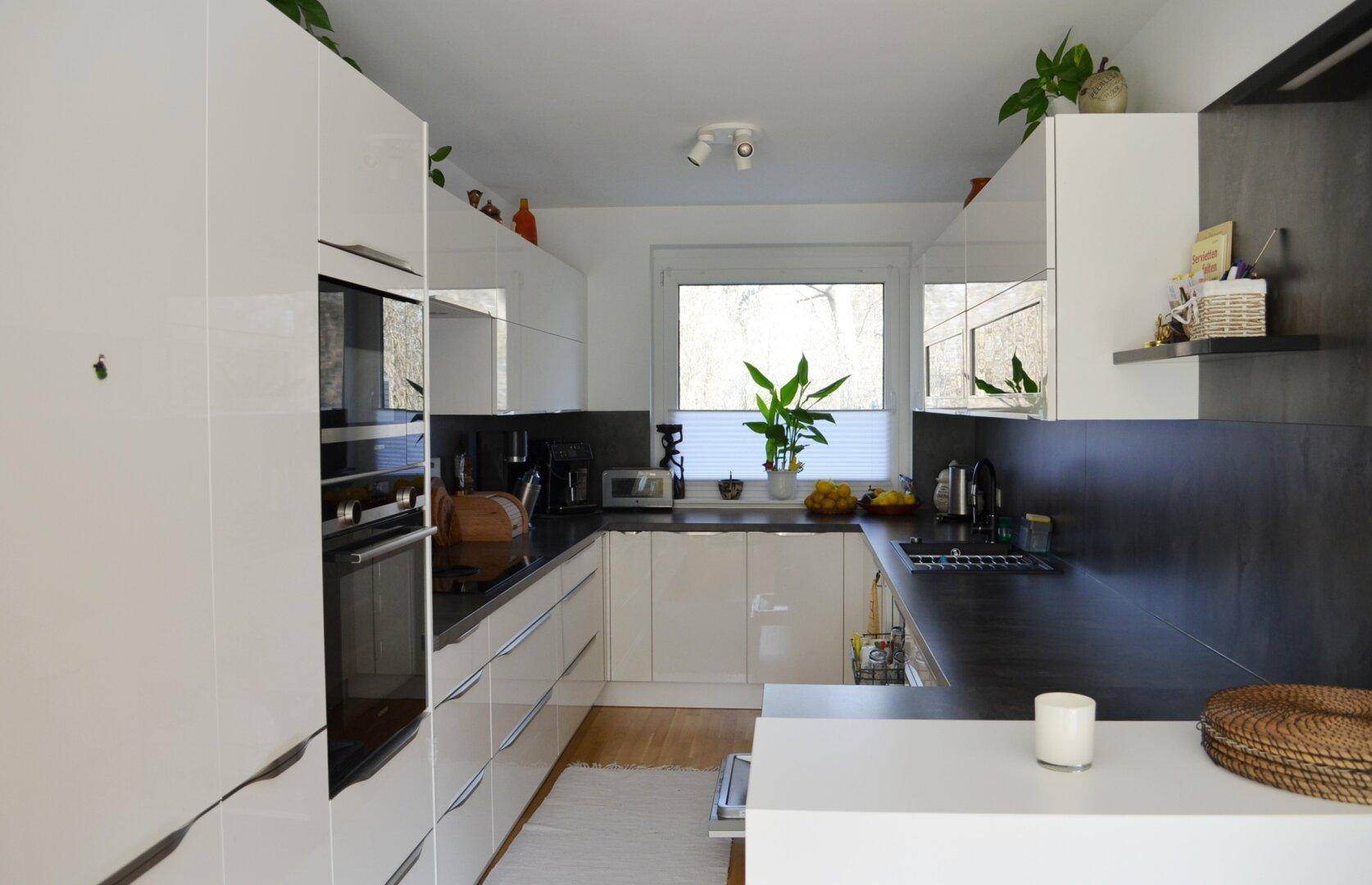 Moderne Küche mit weißen Hochglanzfronten, dunklen Arbeitsplatten und einem Fenster für natürliches Licht.