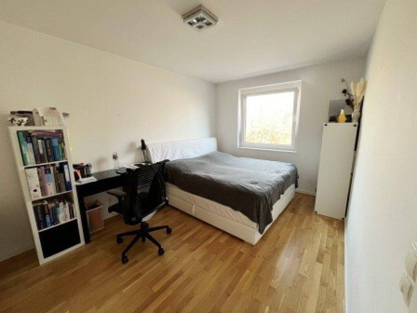 Helles Schlafzimmer mit Holzboden, Bett, Schreibtisch und Bücherregal, ideal als Arbeits- und Ruheraum.