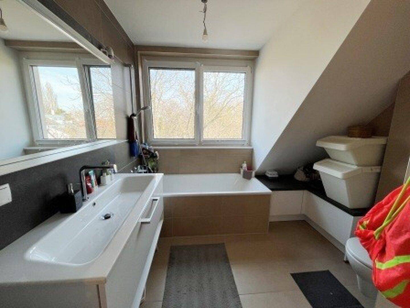 Helles Badezimmer im Dachgeschoss mit langem Waschbecken, Badewanne und Fenster, das viel Licht spendet.