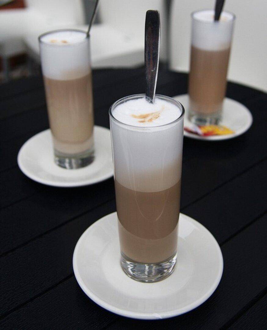 Drei Gläser Latte Macchiato mit Löffeln auf weißen Untertassen auf einem dunklen Tisch.