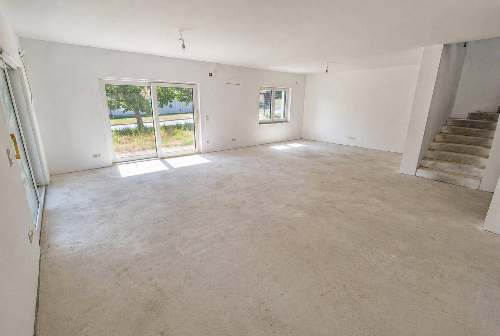 Großer, heller Wohnbereich im Rohbau mit Zugang zur Treppe und viel Tageslicht durch die Fenster.