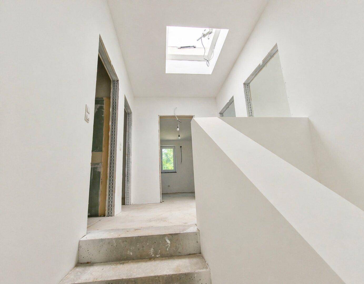 Heller Flur im Rohbau mit Betontreppe und einem Oberlicht, das natürliches Licht spendet.