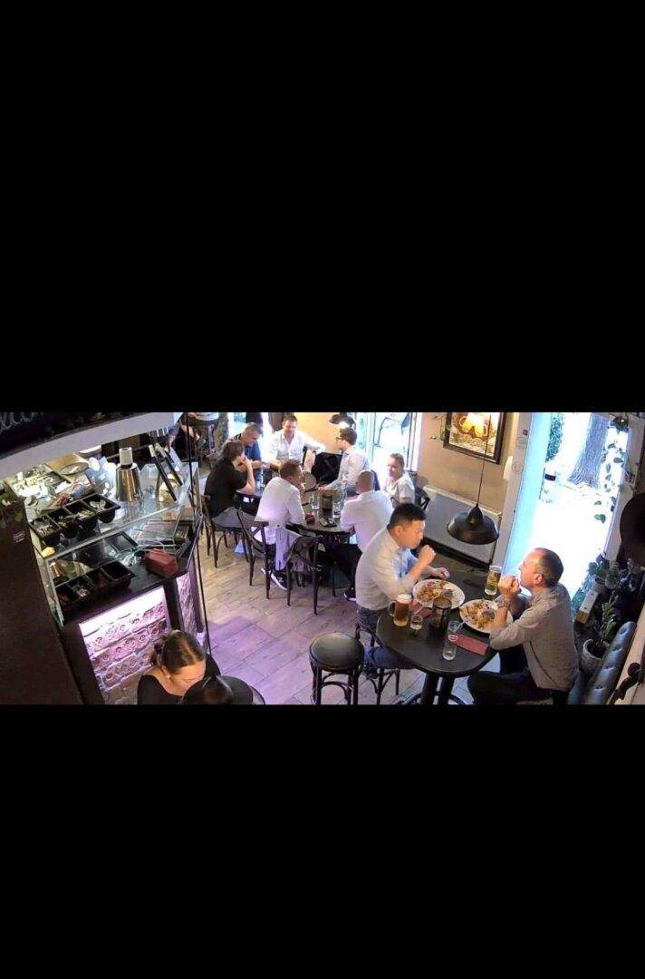 Überwachungsaufnahme eines belebten Restaurantbereichs mit Gästen an Tischen und einer Theke.