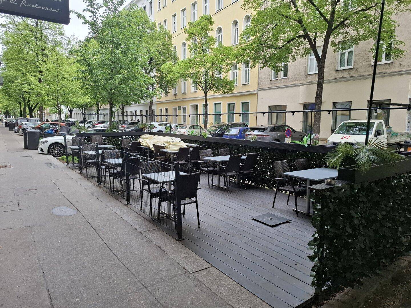 Außenbereich eines Cafés mit Holzterrasse, Tischen und Stühlen entlang einer belebten Straße.