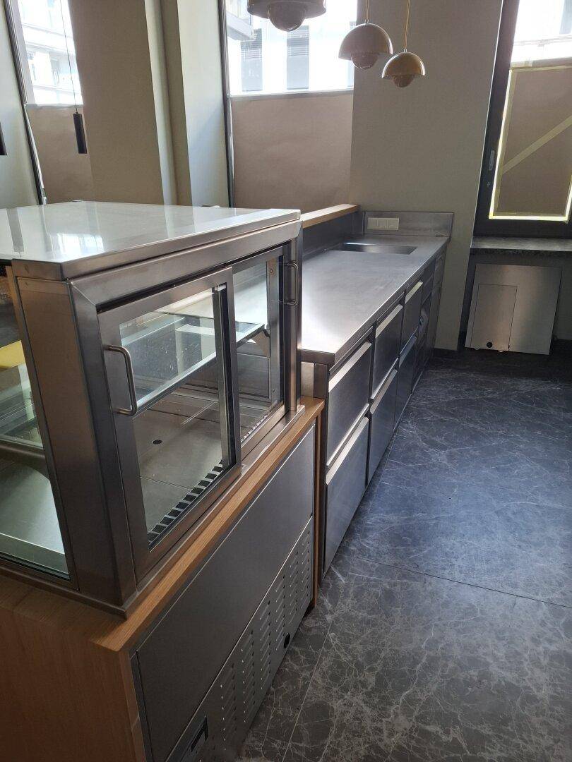 Edelstahl-Arbeitsbereich mit integriertem Spülbecken, Schubladen und einer gekühlten Glasvitrine auf dunklem Marmorboden.