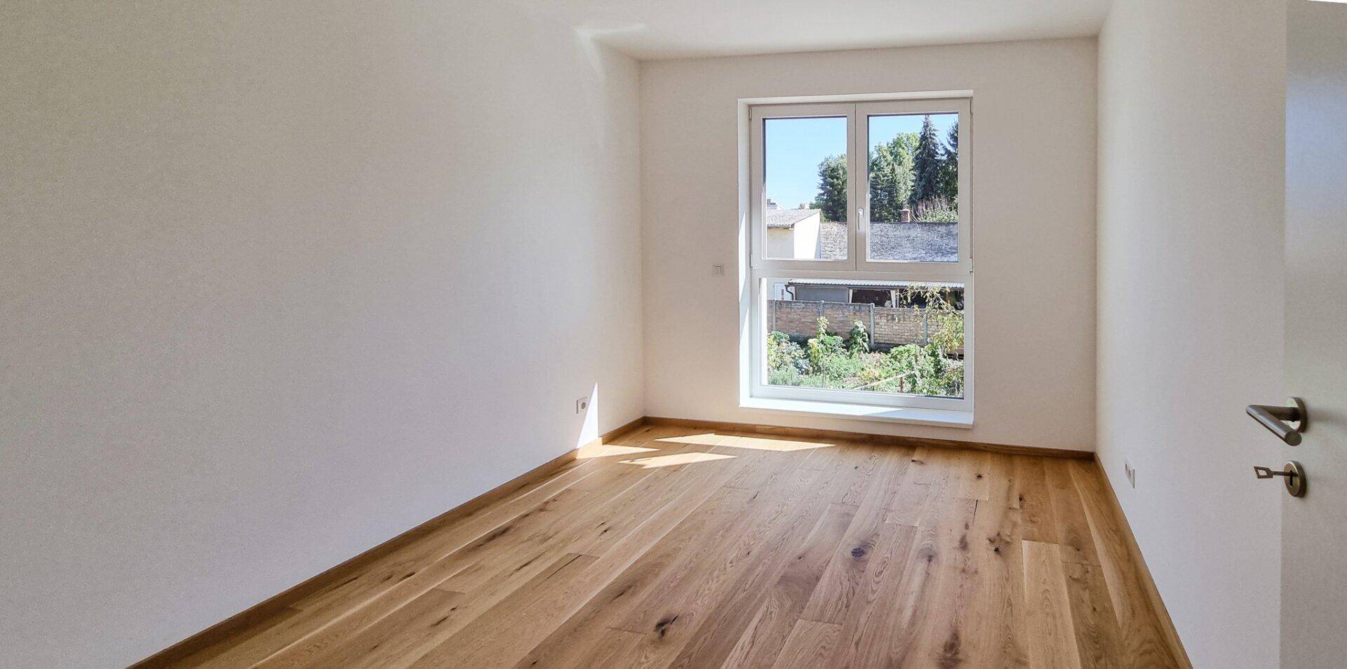 Leerer Raum mit hellem Holzboden und Fenster, das einen Blick in den Garten bietet.