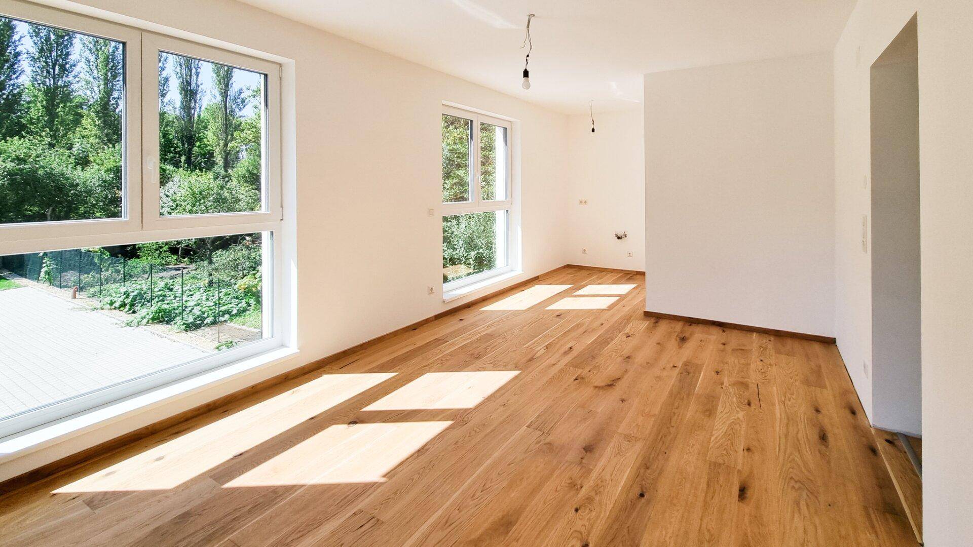 Lichtdurchfluteter, leerer Raum mit Holzboden und großen Fenstern mit Gartenblick.