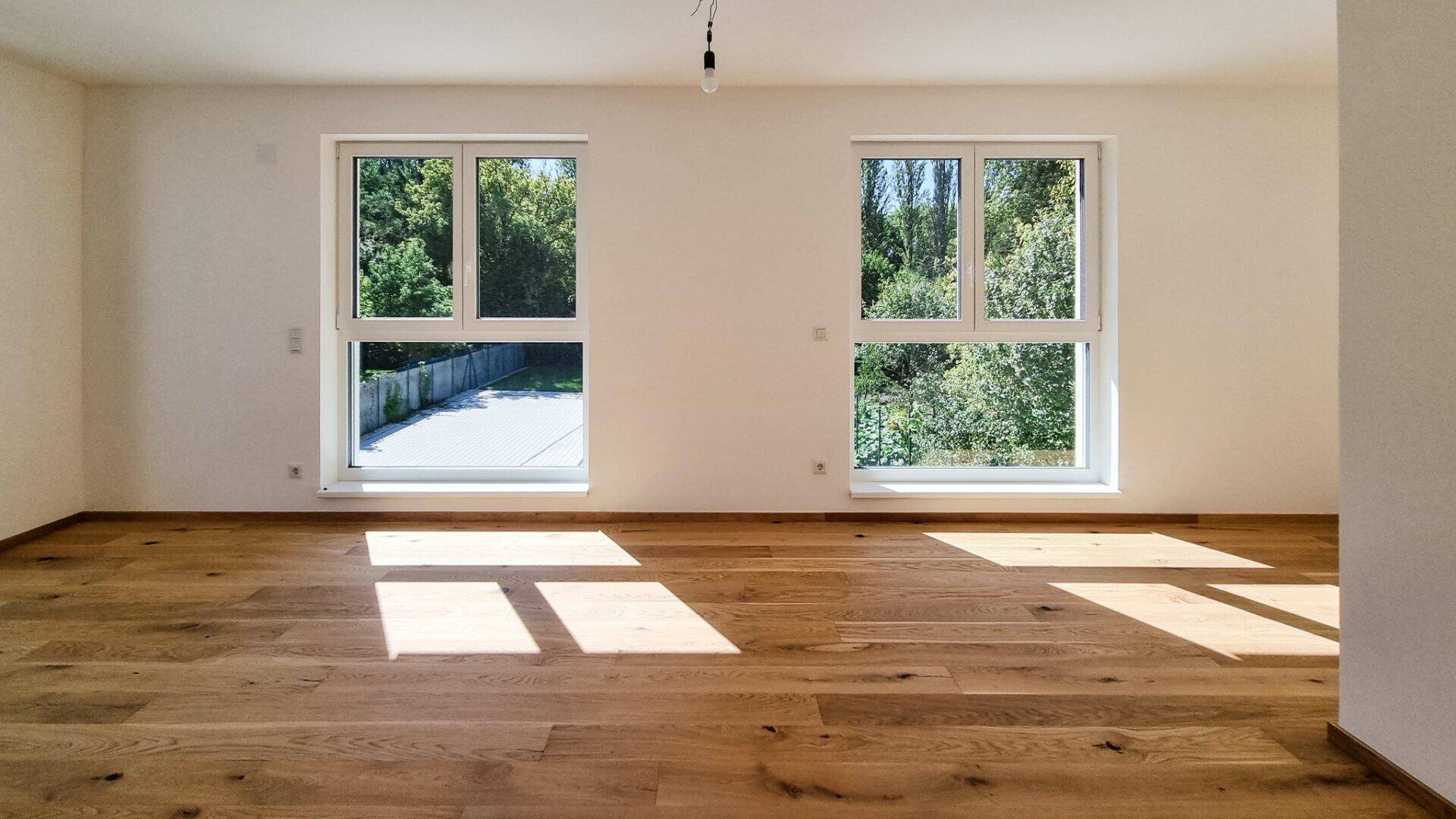 Heller, leerer Raum mit Holzboden und zwei großen Fenstern, die viel Tageslicht hereinlassen.
