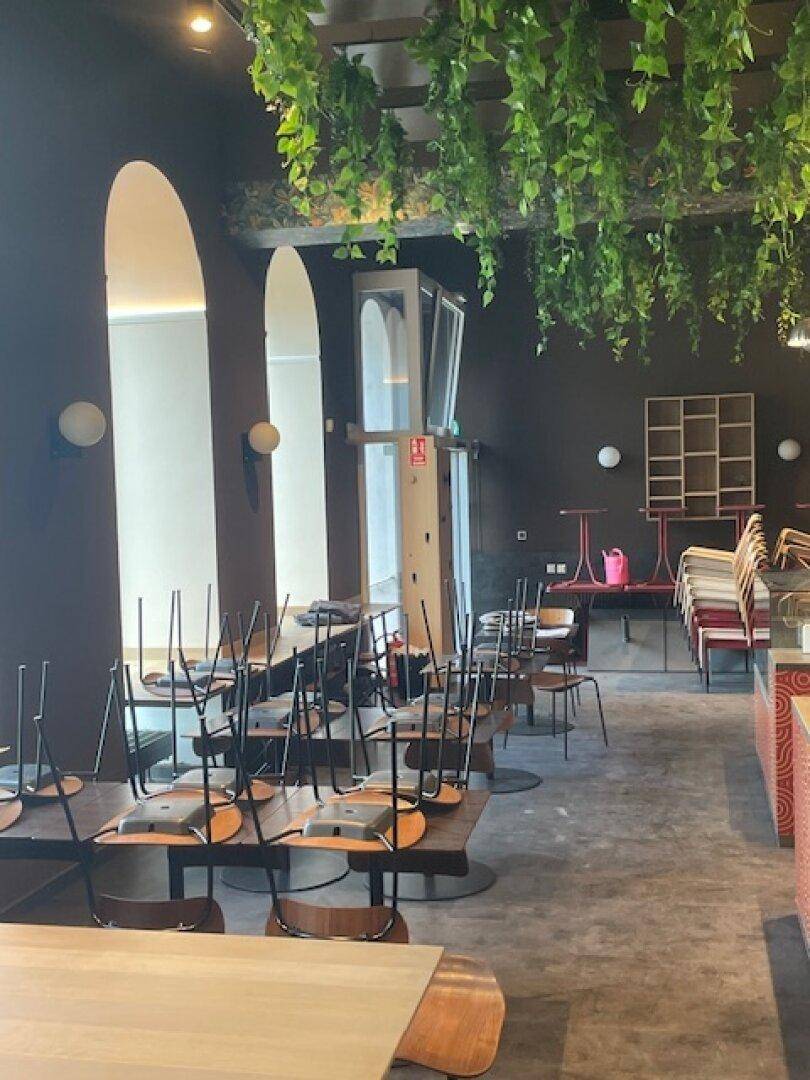 Sitzbereich eines Restaurants mit gestapelten Stühlen, Tischen und großen Bogenfenstern, ein Aufzug ist sichtbar.