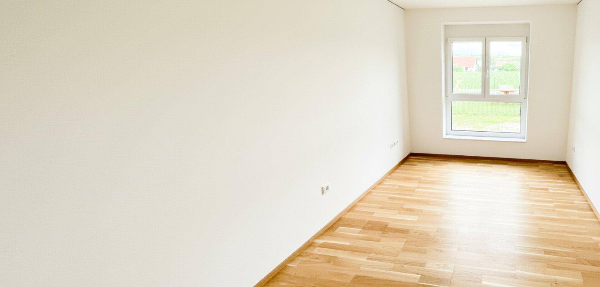 Leeres Schlafzimmer mit hellem Holzboden und einem Fenster, das viel Tageslicht hereinlässt.