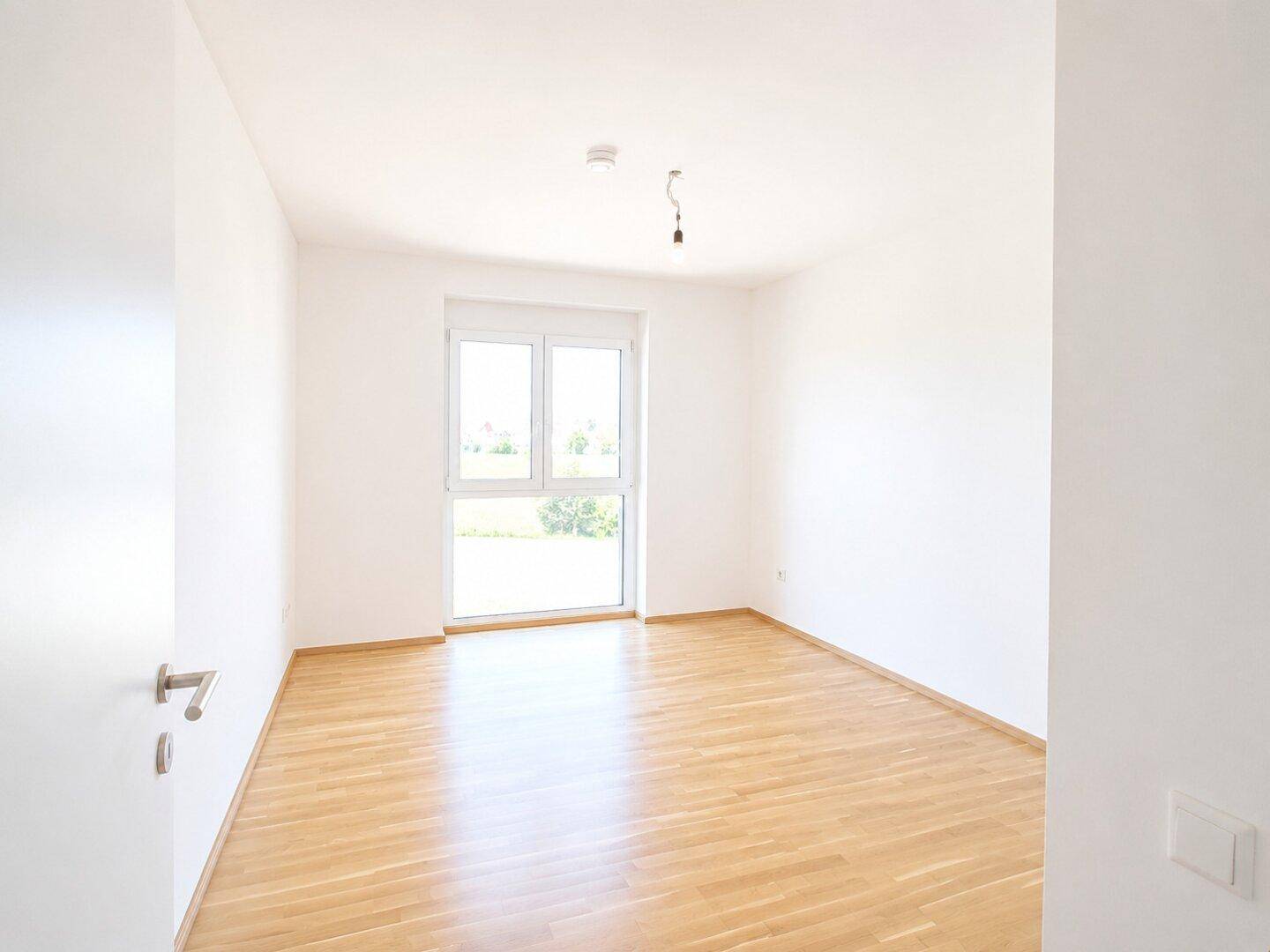 Unmöbliertes Schlafzimmer mit Holzboden und großem Fenster, das viel Tageslicht hereinlässt.