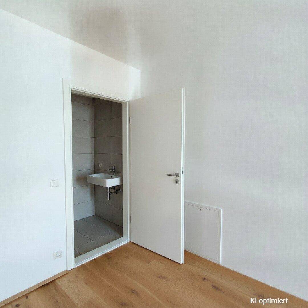Heller Raum mit Holzboden und Zugang zu einem kleinen Bad mit Waschbecken.