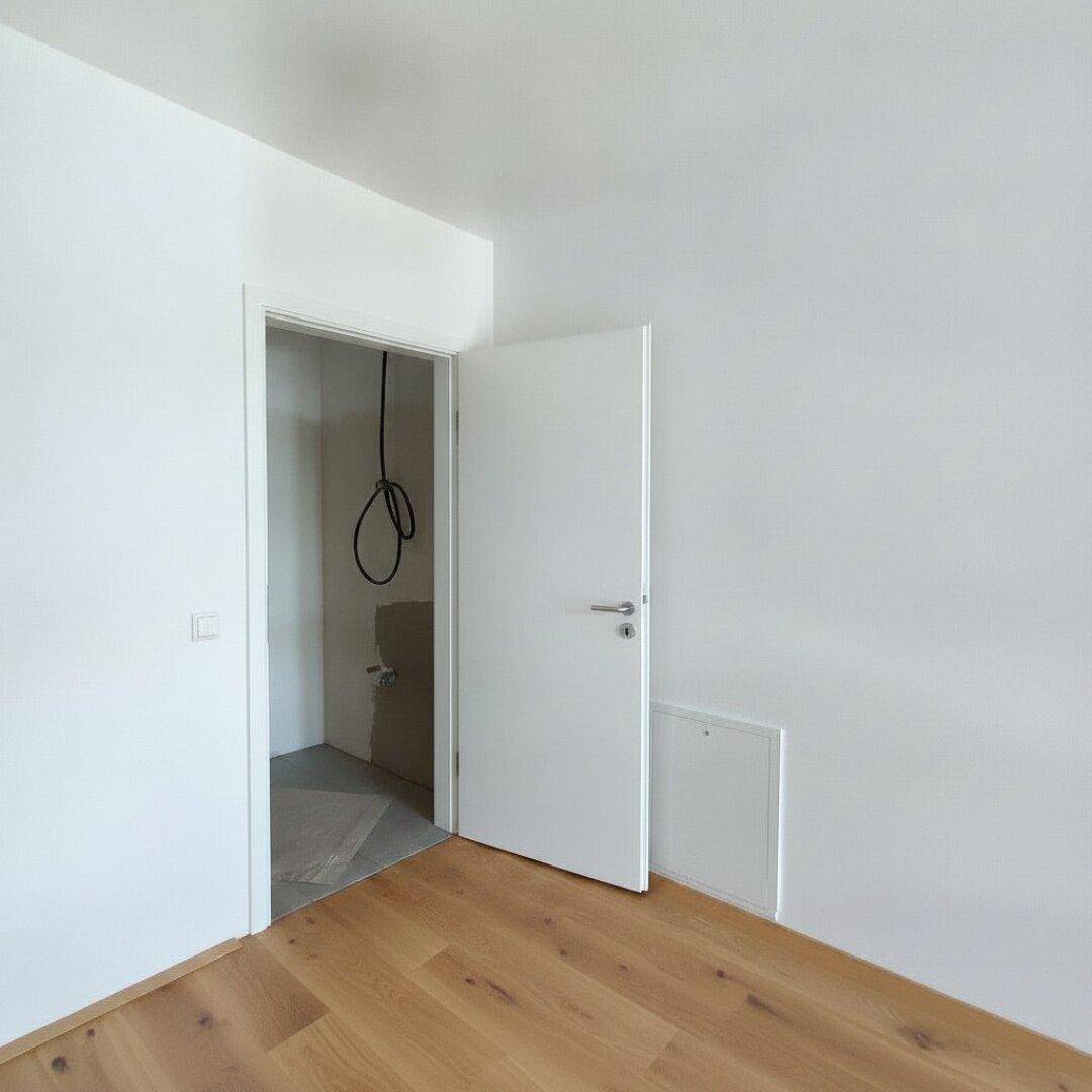 Raum mit Holzboden und offener Tür zu einem unfertigen Bereich mit Installationen.