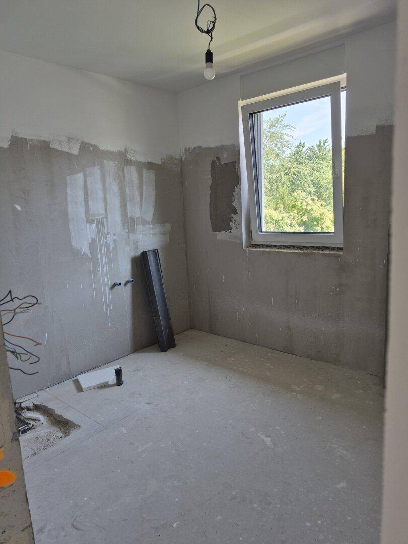 Unfertiges Badezimmer mit Fenster und vorbereiteten Anschlüssen für Sanitärinstallationen.