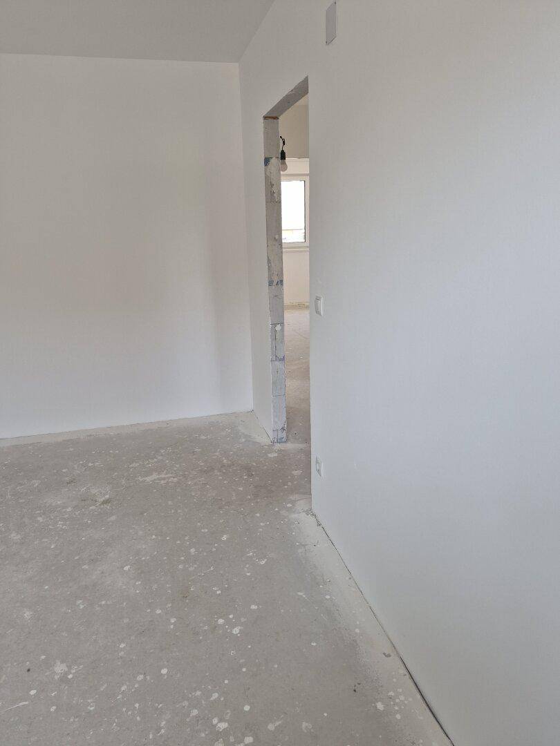 Unfertiger Innenraum mit weiß gestrichenen Wänden und einem Durchgang zu einem weiteren Zimmer.