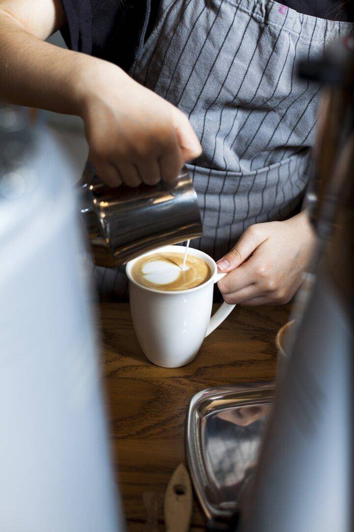 Barista bereitet Latte Art zu, gießt Milch in eine Tasse Kaffee auf einem Holztisch.