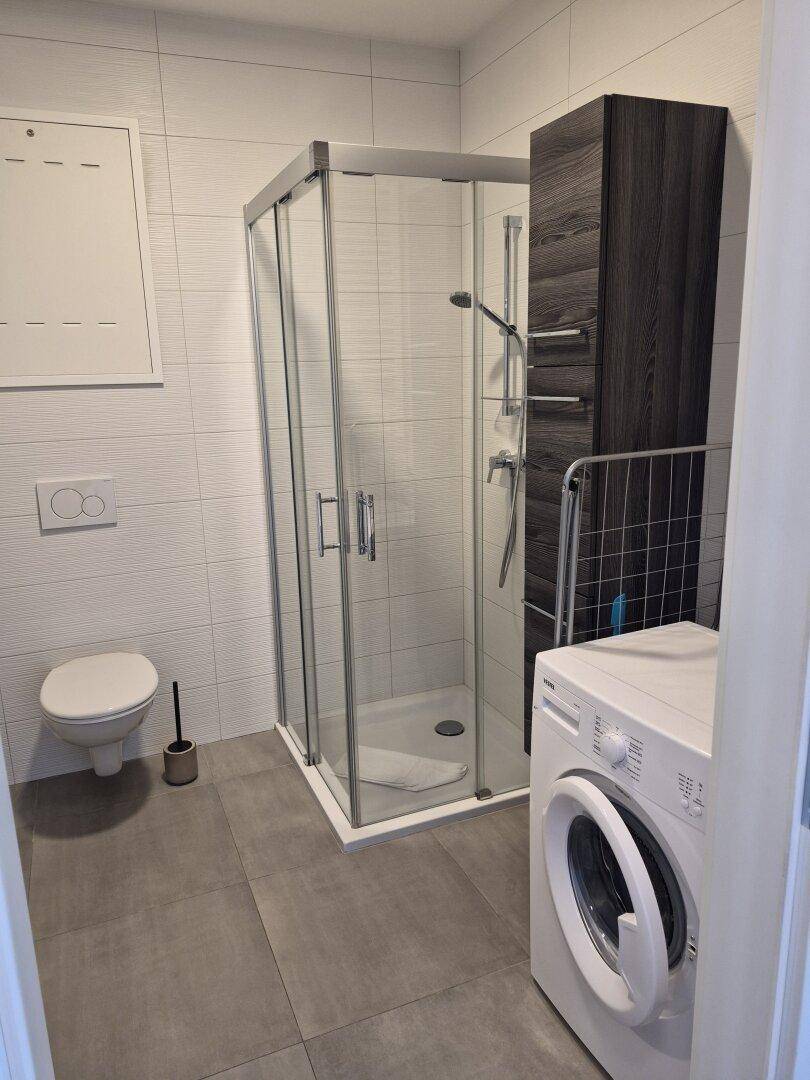 Modernes Badezimmer mit Duschkabine, Wand-WC und Waschmaschine auf grauen Fliesen.