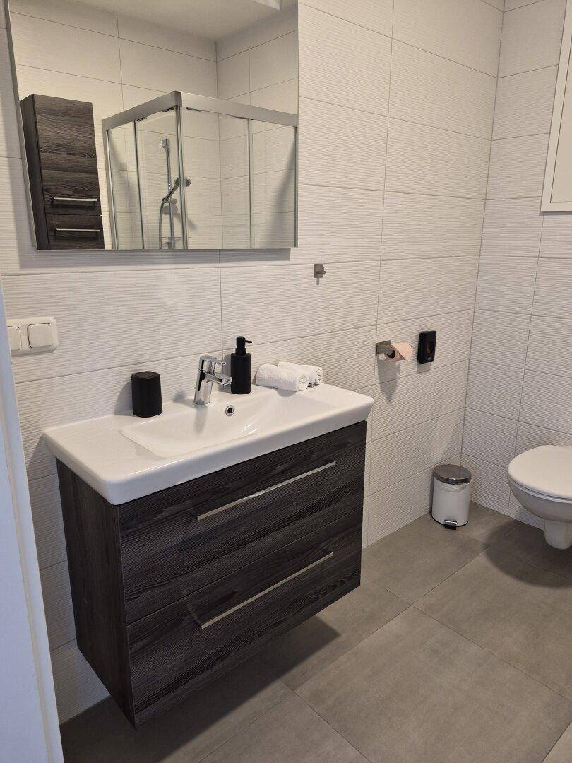 Helles Badezimmer mit modernem Waschtisch, Spiegel und Wand-WC auf grauen Fliesen.
