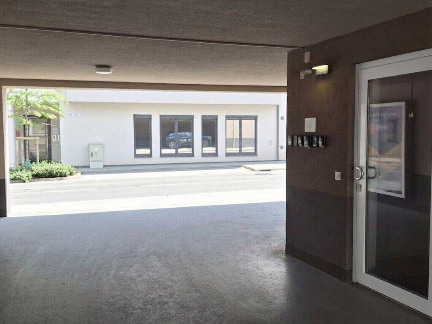 Überdachter Parkbereich vor einem Gebäude mit Eingangstür und Fenstern, der Zugang zum Inneren bietet.