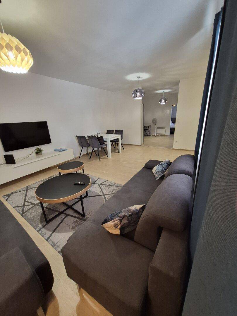 Offener Wohnbereich mit dunklem Sofa, Couchtischen und Essbereich, durch Vorhänge abgetrennt.