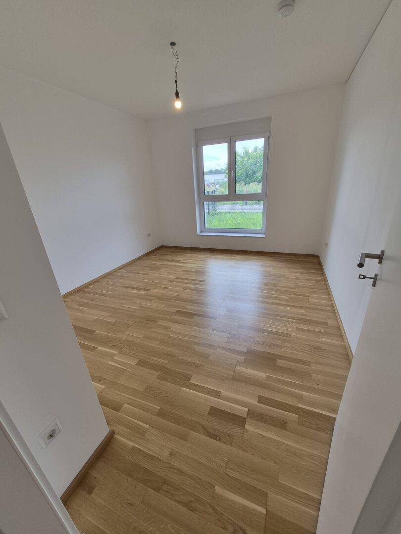 Helles Schlafzimmer mit Parkettboden und einem Fenster mit Blick ins Grüne.