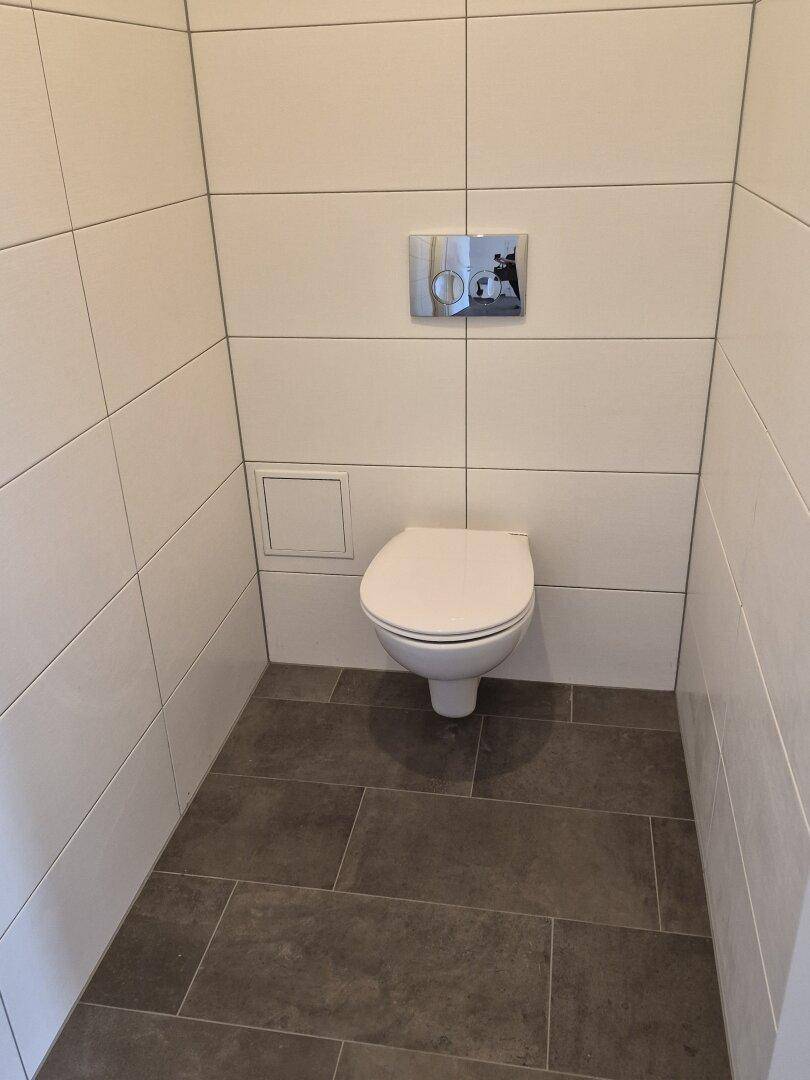 Separates WC mit wandhängender Toilette und weiß-grauen Fliesen an Wänden und Boden.