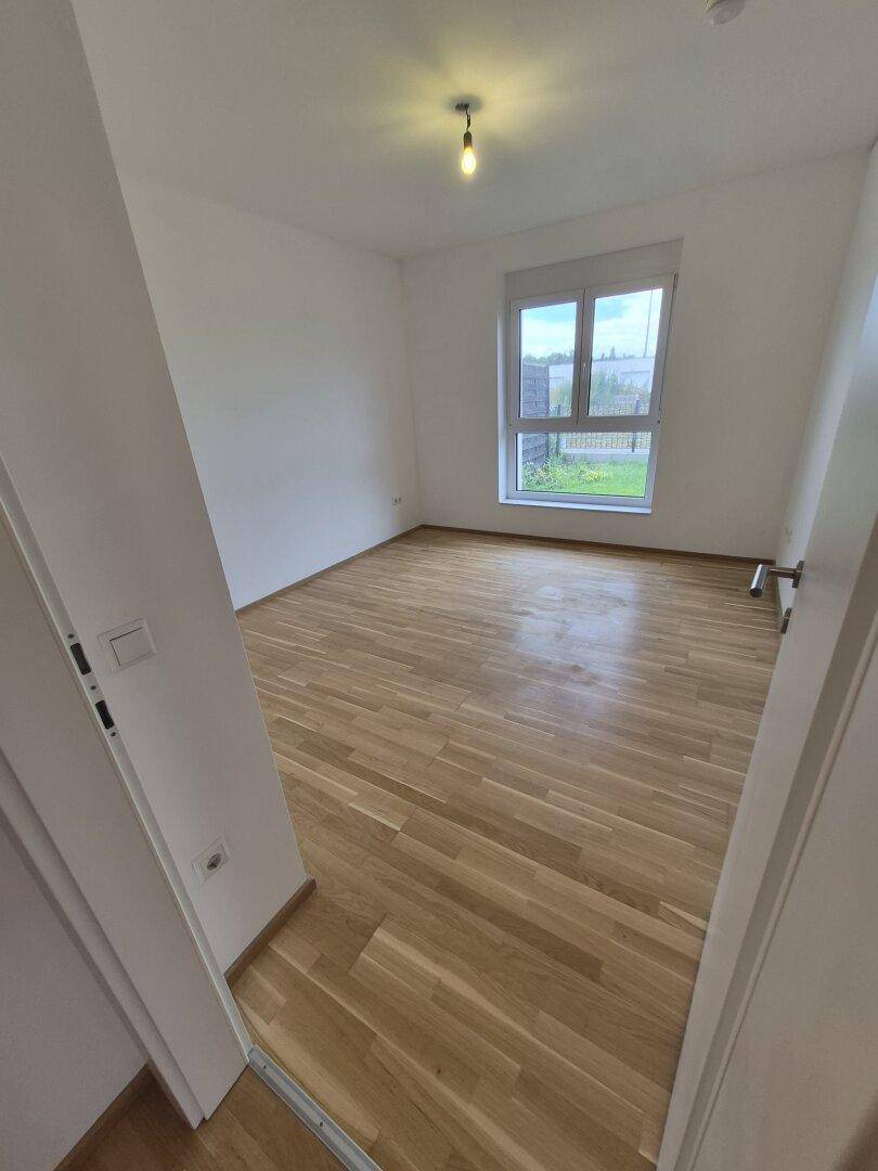 Geräumiges Schlafzimmer mit hellem Holzparkettboden und großem Fenster mit Blick ins Grüne.