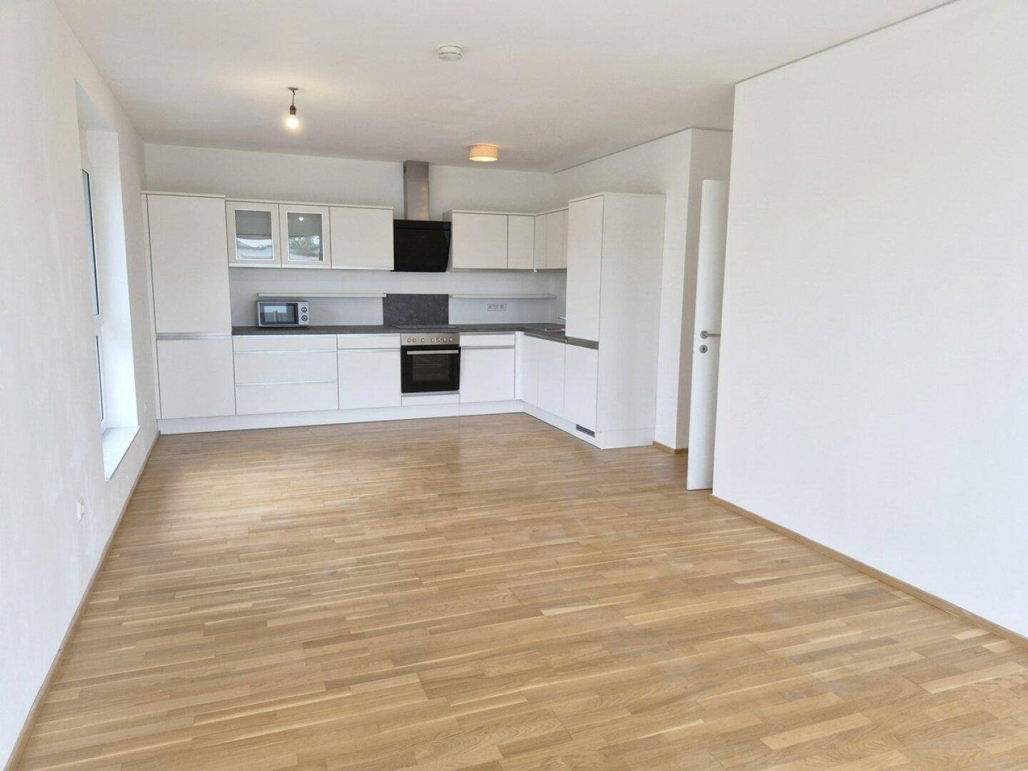 Offener Wohnbereich mit integrierter weißer Küche und durchgehendem Holzparkettboden.