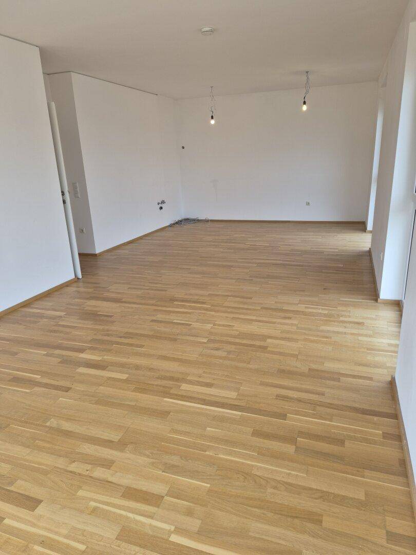 Geräumiges, unmöbliertes Zimmer mit hellem Parkettboden und weißen Wänden.