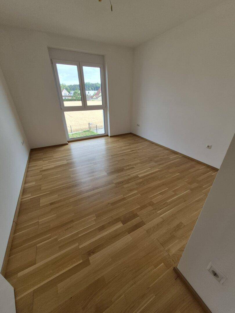 Unmöbliertes Schlafzimmer mit hellem Parkettboden und einem Fenster mit Blick ins Grüne.
