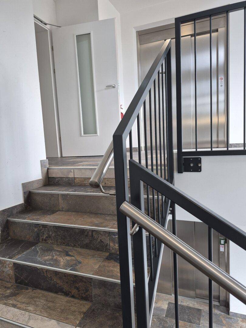 Treppenhaus mit Fliesenstufen und Metallgeländer, daneben befindet sich der Aufzug.
