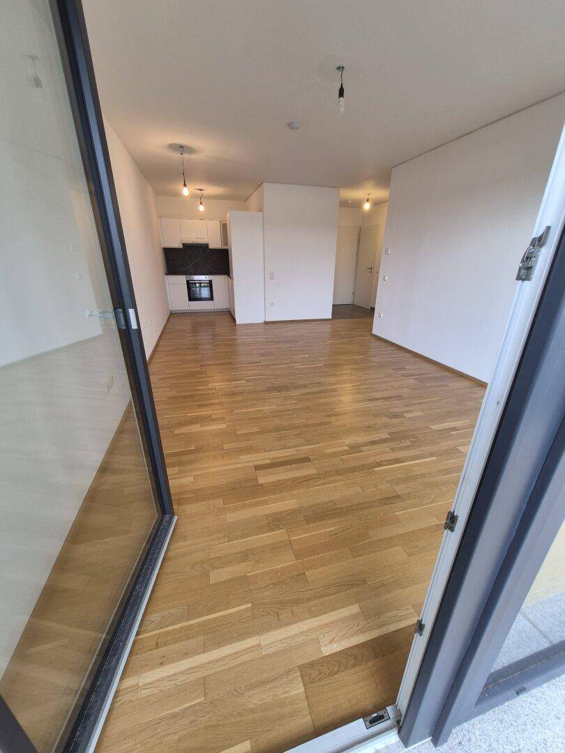 Offener Wohnbereich mit hellem Parkettboden und integrierter Küchenzeile, gesehen durch eine Balkontür.