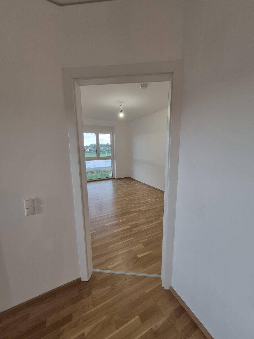 Leeres Zimmer mit hellem Parkettboden und großem Fenster, das viel Tageslicht hereinlässt.