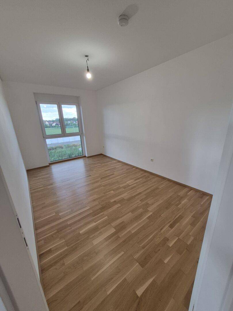 Geräumiges, leeres Zimmer mit Holzboden und Fenster, das einen Blick ins Grüne bietet.