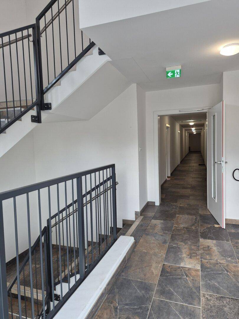 Treppenhaus und langer Flur mit Fliesenboden und Metallgeländer, führt zu den Wohneinheiten.