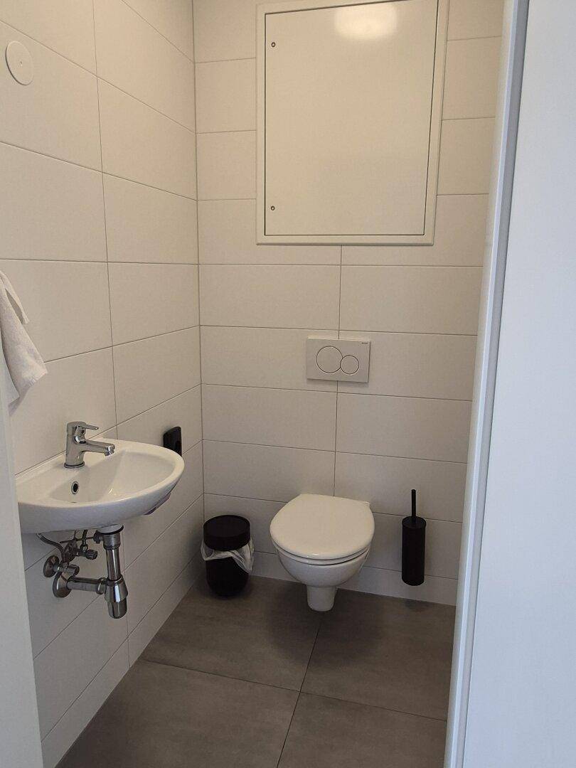 Separates WC mit Wandtoilette, kleinem Waschbecken und weißen Fliesen.