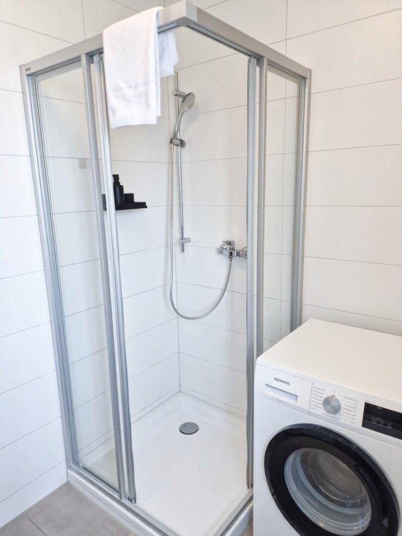 Helles Badezimmer mit Duschkabine, Waschmaschine und weißen Wandfliesen.