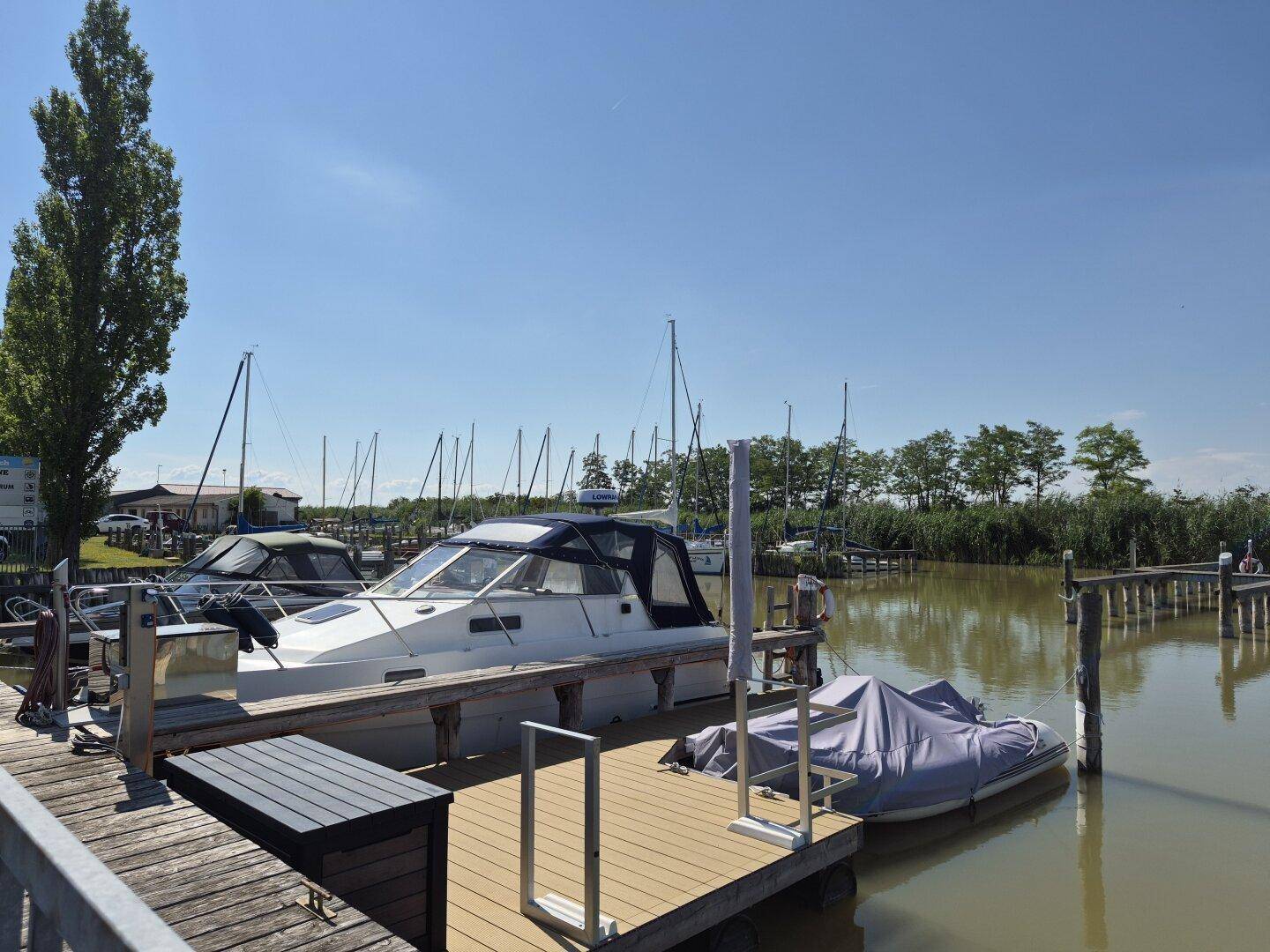 Ein Motorboot liegt an einem Holzsteg in einem Yachthafen mit weiteren Booten im Hintergrund.