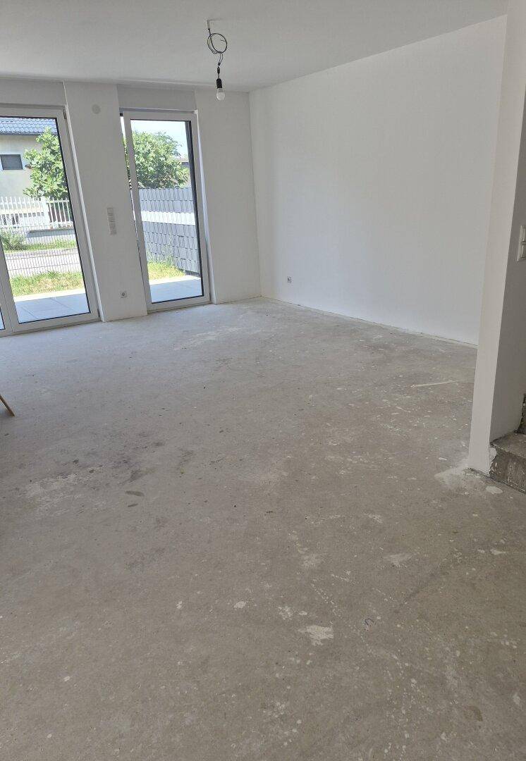 Leerer, unfertiger Wohnraum mit großen Fenstern und Betonboden, bereit für den Ausbau.