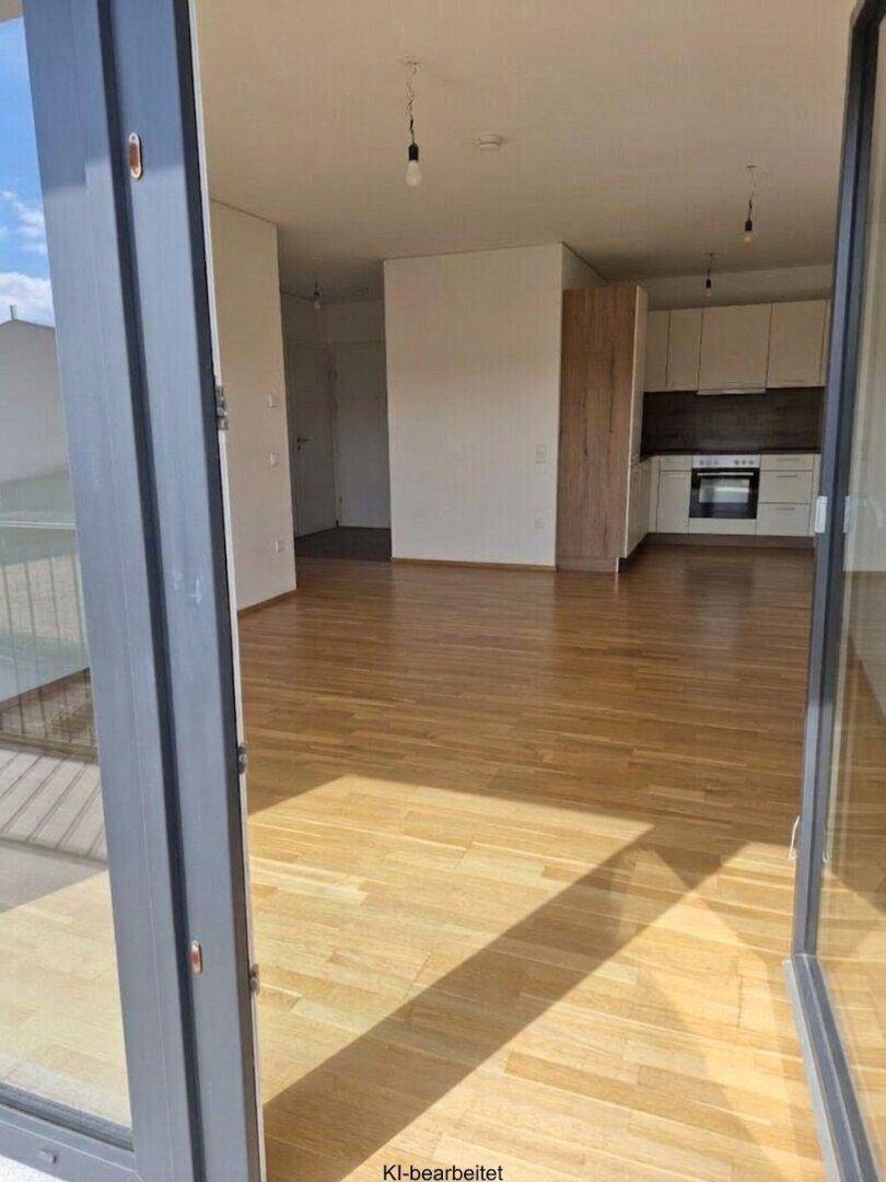 Heller offener Wohnbereich mit Holzboden und Zugang zu einem Balkon, im Hintergrund eine Einbauküche.