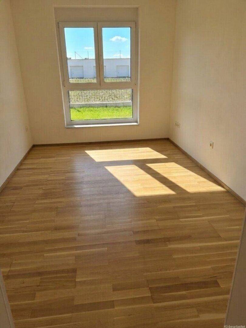 Helles, leeres Zimmer mit Holzparkettboden und großem Fenster mit Blick ins Grüne.