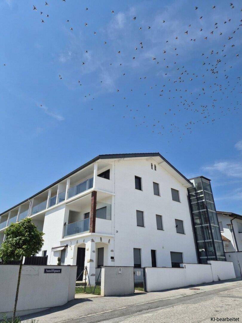 Modernes weißes Gebäude mit Balkonen und einem externen Glasaufzug unter blauem Himmel.