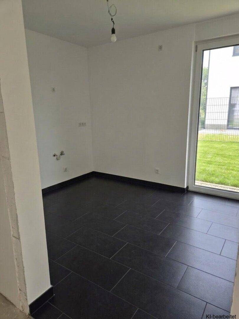 Leerer Raum mit dunklen Fliesen, weißen Wänden und Zugang zum Garten durch eine große Tür.