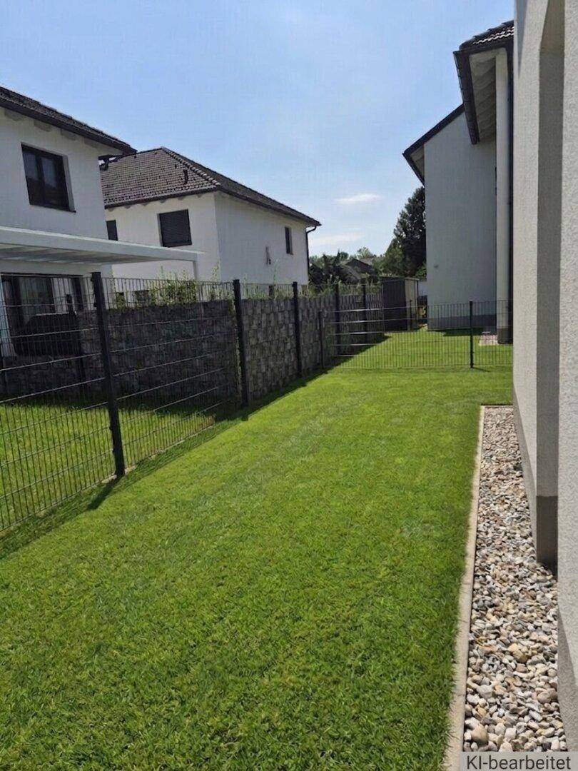 Gepflegter grüner Gartenbereich zwischen zwei Häusern mit Zaun und Kiesstreifen unter der Sonne.