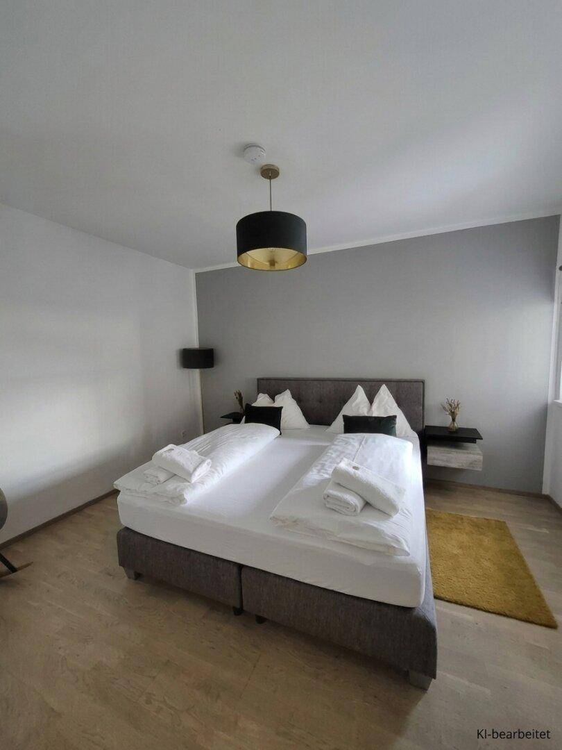 Helles Schlafzimmer mit Doppelbett, Holzboden und moderner Deckenleuchte in Grau- und Weißtönen.