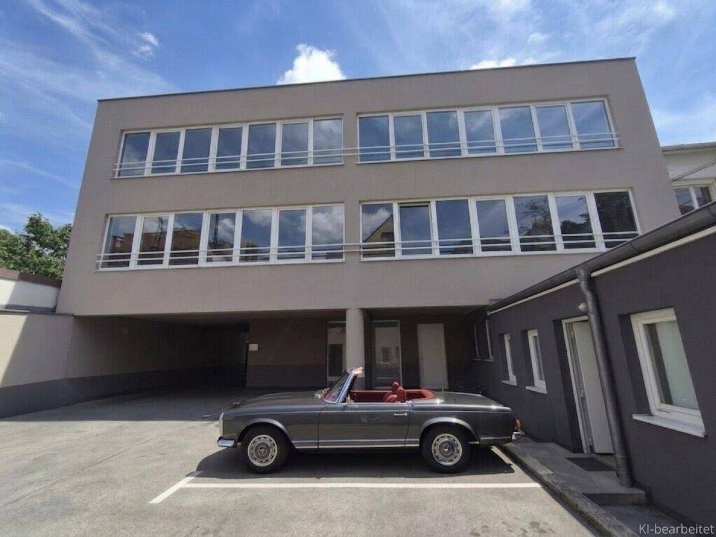 Modernes Gebäude mit großen Fenstern und überdachtem Parkplatz, davor ein klassisches Cabriolet.