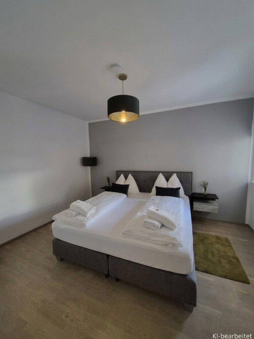 Helles Schlafzimmer mit Doppelbett, Nachttischen und Laminatboden, eine Wand in Grau abgesetzt.