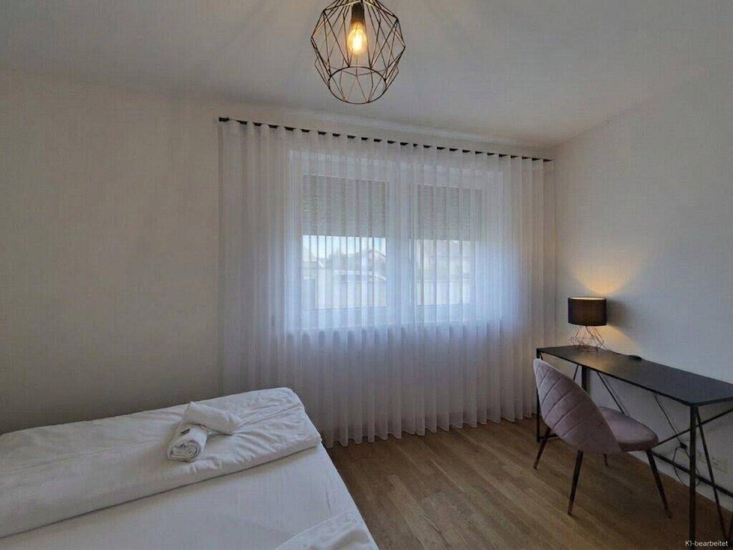 Helles Schlafzimmer mit großem Fenster, Schreibtisch und modernem Pendelleuchten-Design.