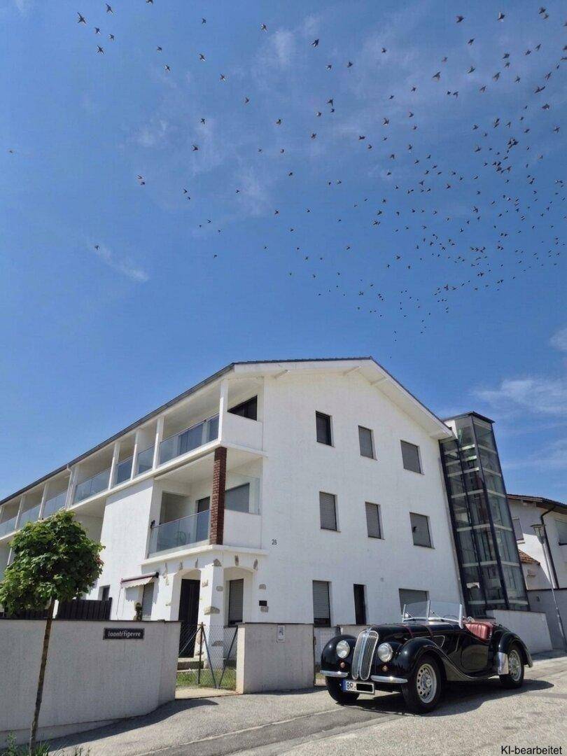 Moderne weiße Gebäudefassade mit Balkonen und einem externen Aufzug unter blauem Himmel.